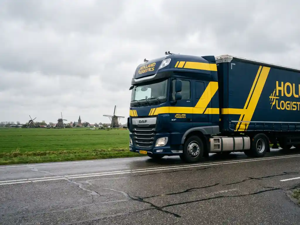 Grote vrachtwagen met donkerblauwe trailer en gele strepen rijdt over Nederlandse snelweg door landschap met molens