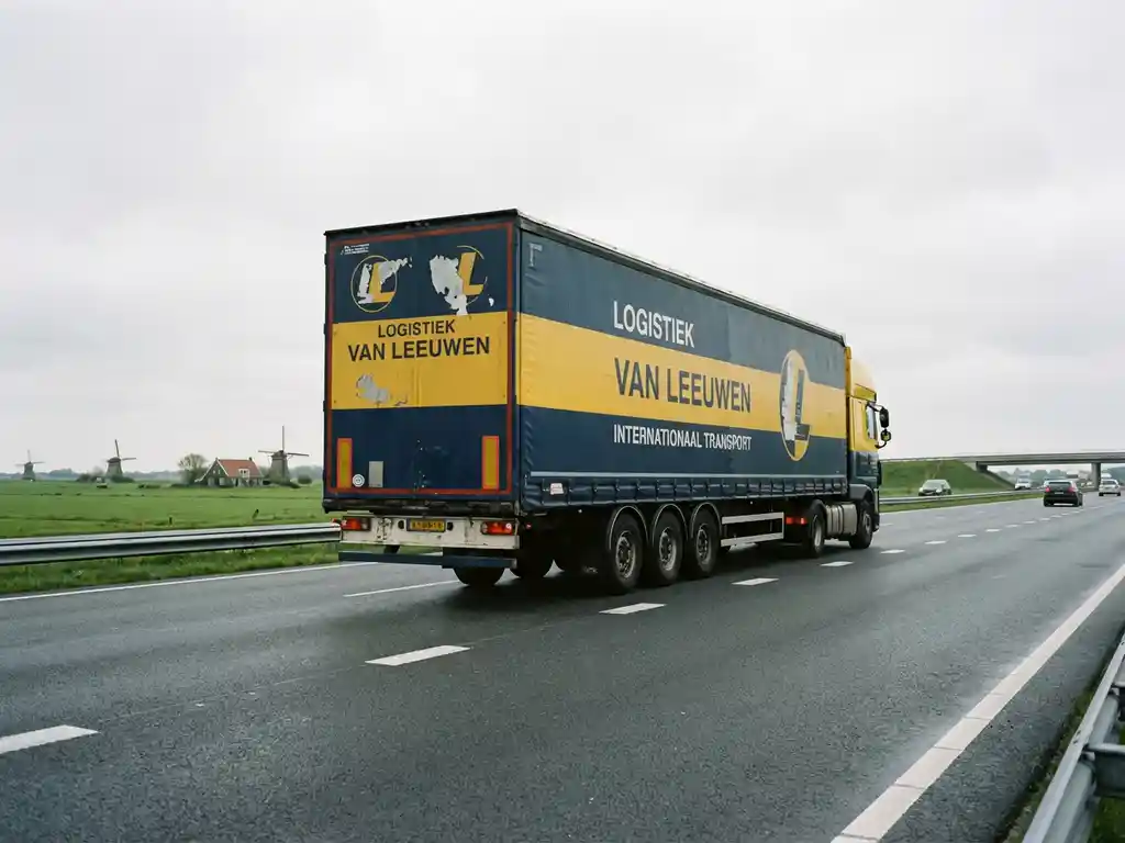 Grote vrachtwagen in marineblauw en geel rijdt over Nederlandse snelweg met groene velden en windmolens op achtergrond