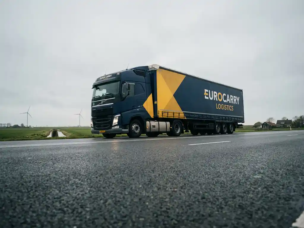 Grote moderne vrachtwagen met donkerblauwe en gele trailer rijdt over Nederlandse snelweg door vlak landschap