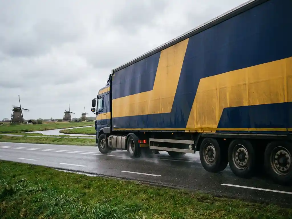 Grote vrachtwagen met marineblauw en goudgeel geometrisch ontwerp rijdt over Nederlandse snelweg met windmolens op achtergrond
