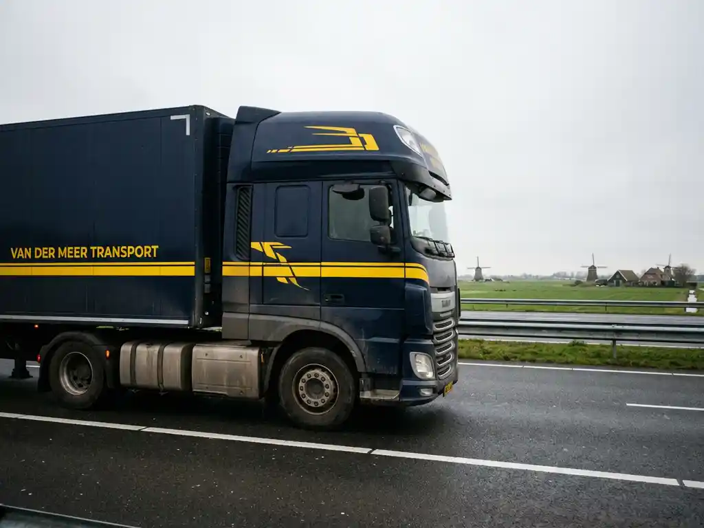 Grote vrachtwagen in marineblauw met gele accenten rijdt over Nederlandse snelweg met groene velden en windmolens op achtergrond