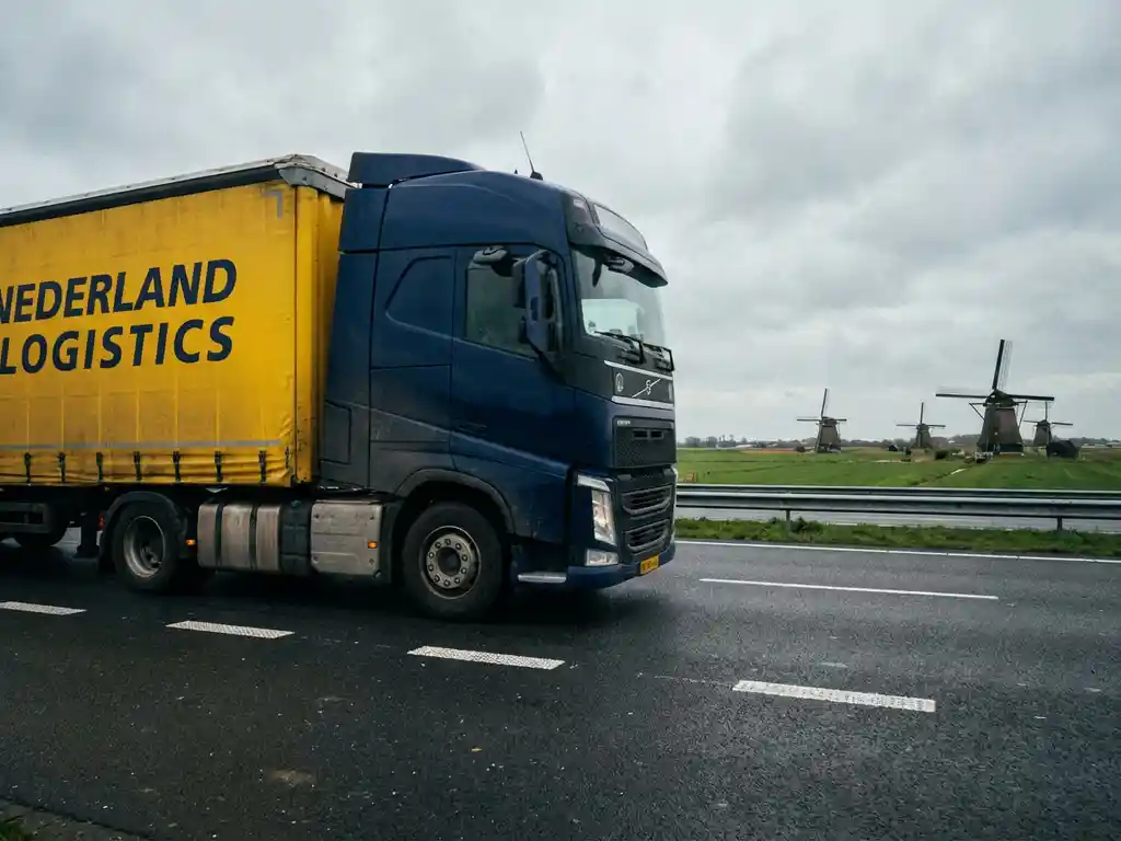 Grote vrachtwagen met donkerblauwe en gele trailer rijdt over Nederlandse snelweg met windmolens op achtergrond